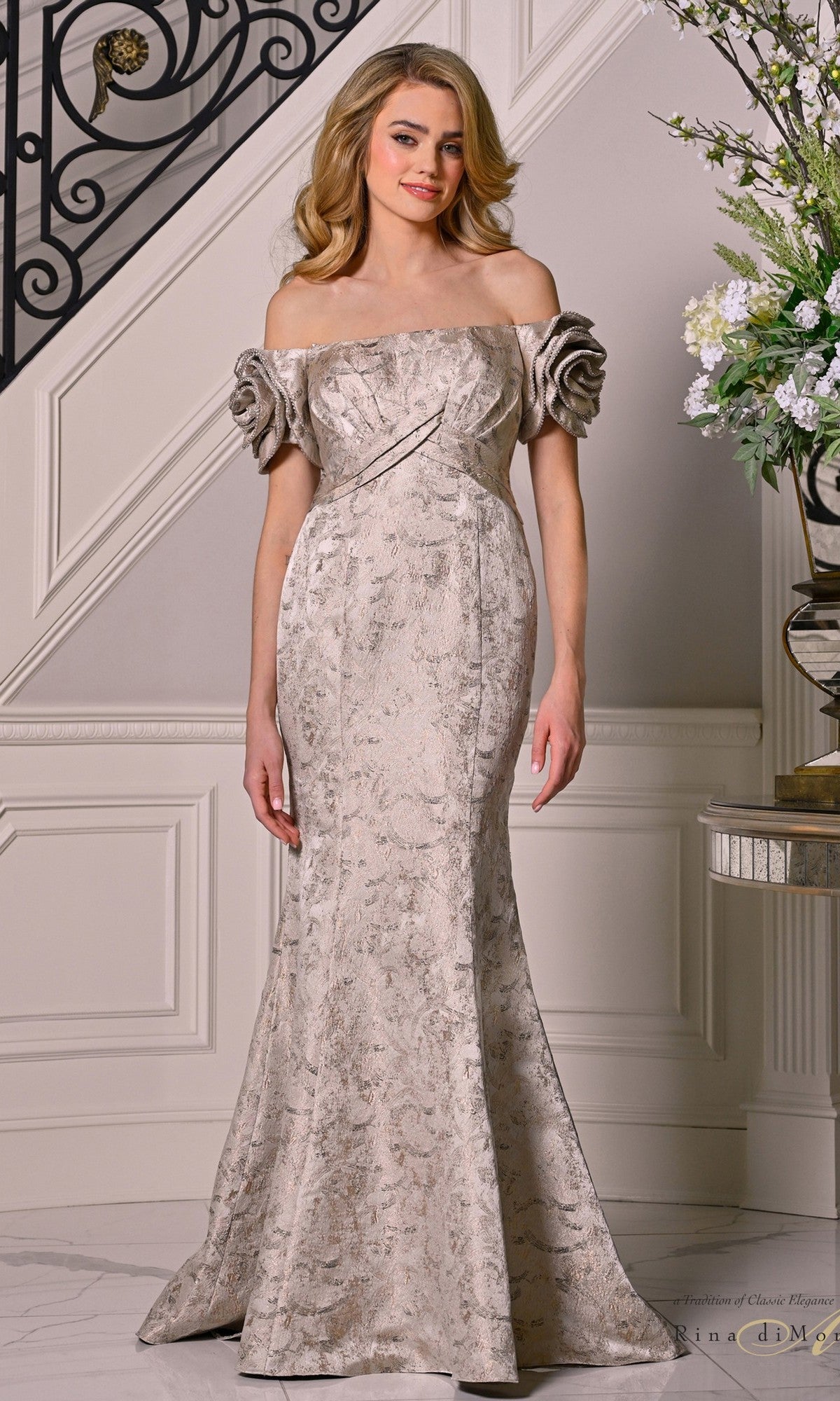 A woman in the Rina Di Montella RD3512 long formal dress—a champagne, off-shoulder gown with rose sleeves and a mermaid silhouette—stands indoors near a staircase and an elegant floral arrangement.
